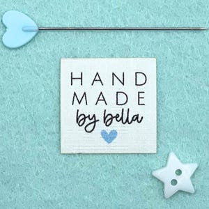 Könnte beinhalten: Ein quadratisches, cremefarbenes Stoffetikett mit den Worten "HAND MADE by bella" in Schwarz und einem blauen Herz. Eine hellblaue herzförmige Nadel und ein weißer sternförmiger Knopf sind ebenfalls auf hellblauem Hintergrund zu sehen.