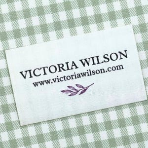 May include: A white rectangular label with the text "VICTORIA WILSON" and "www.victoriawilson.com" in black. A purple leaf design is below the website address. The label is on a green and white checkered background.