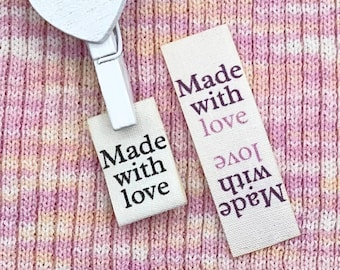 May include: A white heart-shaped clothespin holds a small tag that reads "Made with love" in black text. Another tag with the same text lies on a pink, peach, and white knitted fabric. The tags are rectangular and have a light beige background.