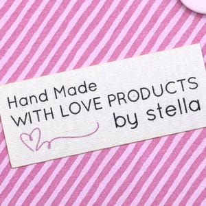 May include: A rectangular, off-white fabric label with black text that reads "Hand Made WITH LOVE PRODUCTS by stella." A pink heart and curved line are embroidered below the words "Hand Made." The background is a pink and white striped pattern.