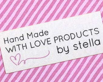 May include: A rectangular, off-white fabric label with black text that reads "Hand Made WITH LOVE PRODUCTS by stella." A pink heart and curved line are embroidered below the words "Hand Made." The background is a pink and white striped pattern.