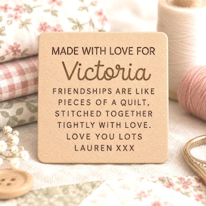 May include: A square, light tan leather tag featuring dark brown engraved text that reads: MADE WITH LOVE FOR Victoria. FRIENDSHIPS ARE LIKE PIECES OF A QUILT, STITCHED TOGETHER TIGHTLY WITH LOVE. LOVE YOU LOTS LAUREN XXX. The tag has rounded corners.
