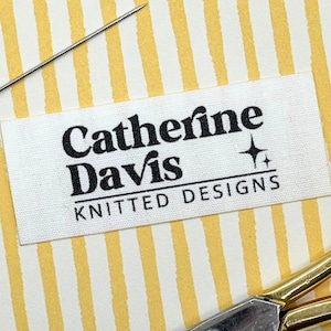 Puede incluir: Una etiqueta de tela blanca con el texto "Catherine Davis KNITTED DESIGNS" en negro. Una aguja y unas tijeras doradas son visibles sobre un fondo a rayas amarillas y blancas. La etiqueta tiene un pequeño gráfico de estrella.