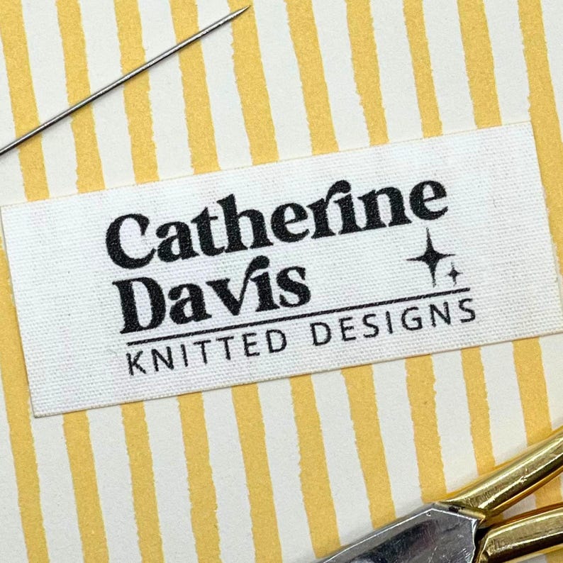 May include: A white fabric label with the text "Catherine Davis KNITTED DESIGNS" in black. A needle and gold scissors are visible on a yellow and white striped background. The label has a small star graphic.