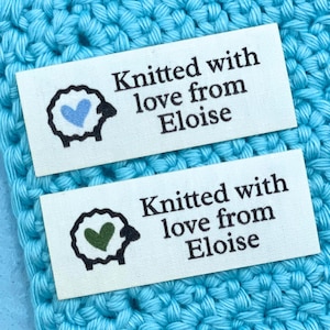 May include: Two rectangular fabric labels with the text "Knitted with love from Eloise" and a sheep illustration with a heart. One sheep has a blue heart, the other has a green heart. The labels are on a blue crocheted surface.