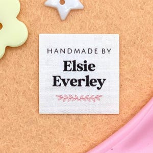 May include: A square, white fabric label with the text "HANDMADE BY Elsie Everley" in black. Below the name is a pink floral design. The label is on a peach-colored surface, with a white star-shaped button and a light green flower-shaped button.