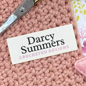 May include: Close-up of a crocheted item in a soft pink hue. A label with the text "Darcy Summers CROCHETED DESIGNS" is visible. Silver and gold scissors are placed diagonally across the top of the crocheted fabric.