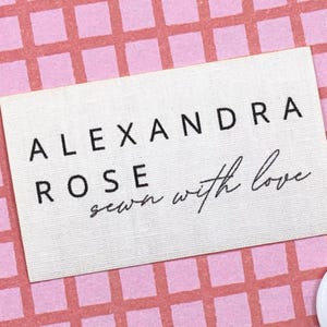 Può includere: Un'etichetta rettangolare bianca con le parole "ALEXANDRA ROSE" in lettere nere. Sotto il nome, la frase "sewn with love" è scritta in corsivo. L'etichetta è posta su uno sfondo rosa con un motivo a griglia.