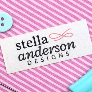 May include: A white rectangular label with the text "stella anderson DESIGNS" in black script. A light pink infinity symbol is above the word "anderson". The label is on a pink and white striped background. A light blue button is in the upper left corner.