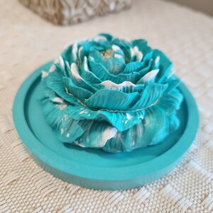 May include: A teal-colored, floral-shaped decorative item. The flower has white accents and gold glitter details, sitting on a matching teal round tray. The item is likely a decorative piece for a home or event.