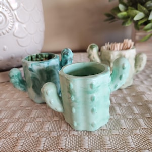 May include: Three ceramic cactus-shaped holders in varying shades of green. The front holder is a light mint green with raised bumps, while the two behind it feature darker marbled green and pale cream finishes, respectively.