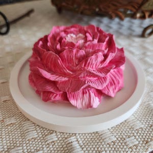 May include: A pink peony flower sculpture with detailed petals sits on a white, round tray. The flower has varying shades of pink and white, creating depth and texture. The tray is simple and elegant, providing a clean backdrop for the floral centerpiece.