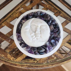 May include: A white, circular decorative bowl with a carved moon face in the center, surrounded by amethyst stones. The bowl sits on a gold-colored, ornate metal surface, creating a celestial and spiritual aesthetic.