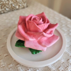 May include: A pink rose sculpture with green leaves sits on a round, off-white tray. The rose is in full bloom, with detailed petals. The tray is on a textured, cream-colored surface, suggesting a decorative item.