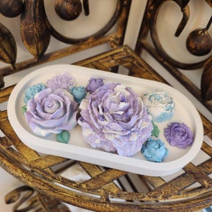 May include: A white, oval decorative tray featuring sculpted, pastel-colored floral arrangements. The flowers include large lavender roses, smaller blue and purple buds, and green leaves. The tray is placed on a gold-colored, ornate metal surface.