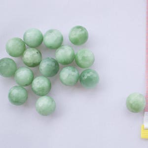 Puede incluir: Una colección de cuentas de piedras preciosas facetadas de color verde pálido. Las cuentas son redondas y tienen un aspecto ligeramente translúcido. Se incluye una cinta métrica para la escala. Las cuentas son probablemente para la fabricación de joyas o manualidades.