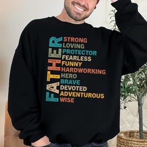 Peut inclure: Sweat-shirt noir avec le mot "FATHER" en turquoise, orange et jaune, avec une liste d'attributs positifs dans une police rétro. Les attributs incluent "STRONG", "LOVING", "PROTECTOR", "FEARLESS", "FUNNY", "HARDWORKING", "HERO", "BRAVE", "DEVOTED", "ADVENTUROUS" et "WISE".