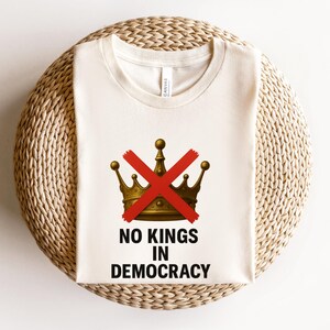May include: Cream-colored t-shirt featuring a gold crown with a red 'X' through it. Below the crown, the text reads 'NO KINGS IN DEMOCRACY' in black. The shirt is displayed on a woven, circular surface.