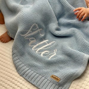 May include: A light blue knitted baby blanket with the name "Walter" embroidered in white script. The blanket has a ribbed border and a small "Monbebe" tag. The blanket is folded and draped over a surface.