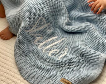 May include: A light blue knitted baby blanket with the name "Walter" embroidered in white script. The blanket has a ribbed border and a small "Monbebe" tag. The blanket is folded and draped over a surface.