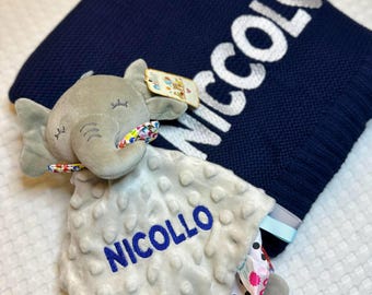 May include: A navy blue knit baby blanket with the name "Niccolo" in white lettering. A gray elephant-shaped security blanket with the name "Niccollo" in blue is in front of the blanket. The security blanket has a textured surface and colorful ribbons.