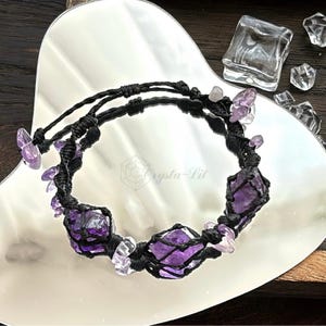 May include: A handmade bracelet featuring raw amethyst stones and smaller clear crystals, strung on black macrame cord. The bracelet is displayed on a reflective surface. The brand name "Crysta-Lit" is visible.