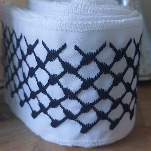 Puede incluir: Un rollo de tela blanca con un patrón geométrico negro. El patrón es un diseño de red repetitivo. La tela está enrollada, mostrando múltiples capas. El fondo está borroso, con una superficie de madera en primer plano.