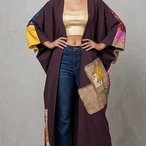 May include: A long, dark purple kimono-style robe with colorful patchwork accents on the sleeves and lower front. The robe is worn over a gold crop top and blue jeans. The sleeves are wide and the robe is open in the front.