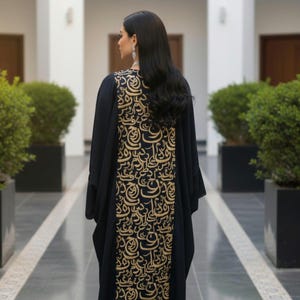 Könnte beinhalten: Schwarze Abaya mit goldfarbenem arabischen Kalligraphie-Design auf dem Rücken. Das langärmlige Kleidungsstück wird von einer Person mit langen schwarzen Haaren getragen. Die Abaya ist bodenlang und hat eine lockere Passform.