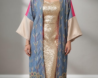 Op de afbeelding: Een vrouw draagt een gouden paillettenjurk en een lange, open blauwe geborduurde jas. De jas heeft roze en beige accenten en ingewikkelde bloemen- en geometrische patronen, wat een elegante outfit creëert.