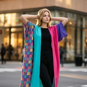 May include: A colorful, patterned kimono-style garment with a vibrant mix of colors. The garment features a patchwork design on one side, a turquoise panel in the center, and pink and purple sleeves. The person is wearing a black top and pants.