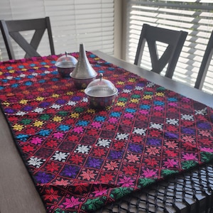 Puede incluir: Un camino de mesa rectangular bordado con un fondo rojo oscuro y un patrón floral repetido en varios colores. El camino de mesa tiene una franja negra. Tres recipientes de metal plateado están sobre el camino de mesa.