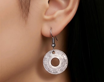 May include: Silver coin earrings with a circular design and a central hole. The earrings are suspended from a simple hook. The coin has intricate details and text around the edge.