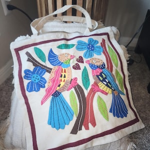 May include: Cream-coloured tote bag with a vibrant embroidered design. The design shows two birds on a branch with blue flowers and green leaves. The bag has a maroon border and cream-coloured handles.