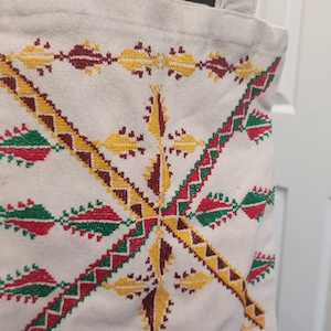 May include: Cream tote bag with a geometric embroidered design in red, yellow, and green. The bag has a simple, rectangular shape with sturdy, off-white straps. The embroidered pattern is symmetrical and features repeating shapes.