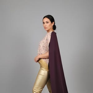 May include: A woman wearing a formal outfit. The ensemble includes gold pants, a patterned top with fringe, and a long, dark purple cape. The outfit is completed with gold high heels.
