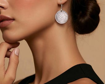 May include: Silver coin earrings with a detailed design. The round earrings hang from silver hooks. The coin design features a central emblem surrounded by text. The earrings are worn by a person in a black top.