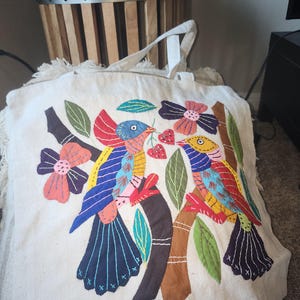 May include: Off-white tote bag with a colorful appliqué design featuring two birds perched on a tree branch. The birds are blue, yellow, and red, with pink and purple flowers and green leaves. The bag has fringe along the bottom.