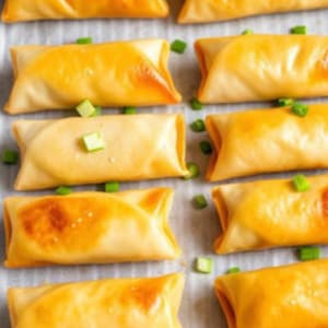 Może przedstawiać: Zbliżenie na blachę do pieczenia wypełnioną złocistobrązowymi, prostokątnymi ciastami, prawdopodobnie sajgonkami. Każde ciasto jest udekorowane małymi zielonymi kawałkami, prawdopodobnie szczypiorkiem. Ciasta mają lekko spęcherzony wygląd, co sugeruje, że są świeżo upieczone.