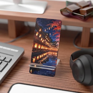 May include: A phone stand with a vibrant illustration of a Japanese street scene at night. The artwork depicts illuminated lanterns reflecting in the water, with colorful trees and buildings. The stand is white and holds a phone or tablet.