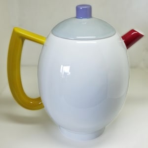 Może przedstawiać: Biały ceramiczny czajnik z żółtym uchwytem, jasnoniebieską pokrywką, fioletowym pokrętłem i czerwonym dzióbkiem.