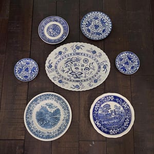 May include: A collection of blue and white porcelain plates and an oval platter. The plates feature various patterns, including floral designs and scenic landscapes. The arrangement is set against a dark wood background, showcasing the classic tableware.