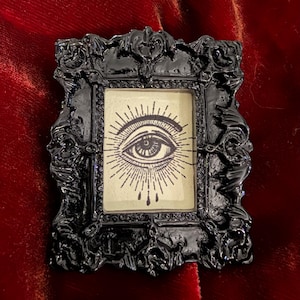 May include: A small rectangular black frame with ornate, raised scrollwork detailing. Inside the frame is a cream-colored rectangular insert featuring a black ink illustration of a single eye with radiating sunburst lines and three teardrop shapes.