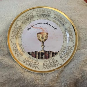 May include: A decorative plate with a gold rim and ornate floral patterns. The center features a design of a chalice above a stack of books, with the text "The faerie wine made me do it!"