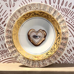 May include: A stack of decorative plates with gold and white accents. The top plate features a heart-shaped design with a blue eye and pearl embellishments. The plates have ornate gold rims and a vintage aesthetic.