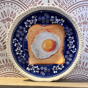 May include: A round, blue and white decorative plate with a fried egg on toast design. The plate has a scalloped edge and a floral pattern. The egg yolk is a bright yellow, and the toast is golden brown.