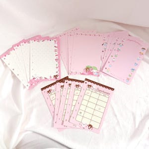 May include: Pink Sugarbunnies stationery set. Includes pink notepaper with decorative borders, lined pages, and calendar sheets. The stationery features Sugarbunnies characters and illustrations of cakes and desserts. The set is arranged on a white surface.