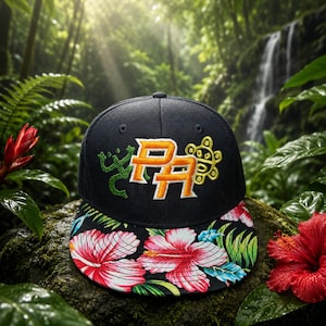 May include: A black baseball cap featuring an embroidered orange and white PA logo on the front, flanked by a green lizard and a yellow sun symbol. The flat brim displays a vibrant floral pattern with large pink hibiscus flowers and green leaves.