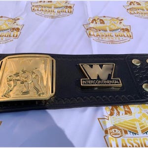 May include: A black leather wrestling championship belt with gold-colored plates. The center plate features a wrestling scene, and the belt has the text "Intercontinental" and the letter "W". The background has a repeating logo.