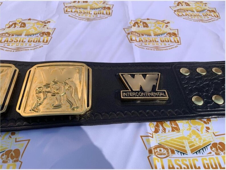 May include: A black leather wrestling championship belt with gold-colored plates. The center plate features a wrestling scene, and the belt has the text "Intercontinental" and the letter "W". The background has a repeating logo.